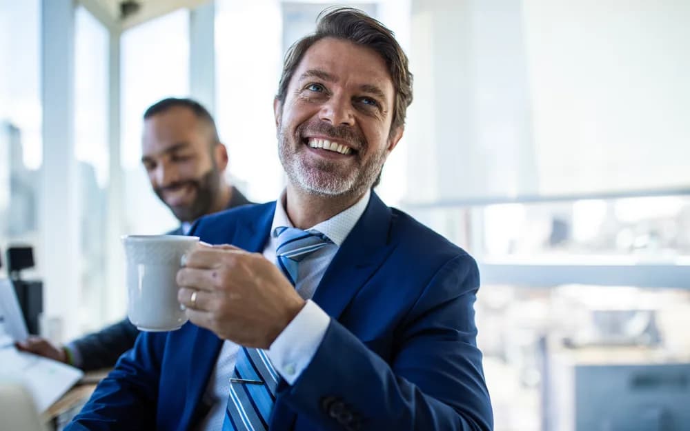 Selbstbewusste Führungskraft im blauen Anzug lächelt in einem modernen Büro mit Skyline-Blick – Symbolbild für inverses Headhunting und den verdeckten Stellenmarkt